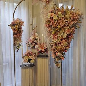 An elegant floral display featuring two interlocking gold metal arches. A large, cascading arrangement of peach lilies, white roses, and greenery hangs from the right side, while a smaller floral cluster sits on the left. In the foreground, white fluted pedestals hold additional flower arrangements and lit taper candles. Luxe Double Arch & Fluted Plinth Set