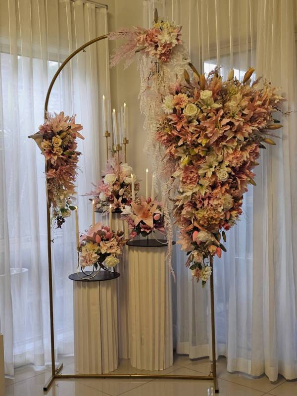 An elegant floral display featuring two interlocking gold metal arches. A large, cascading arrangement of peach lilies, white roses, and greenery hangs from the right side, while a smaller floral cluster sits on the left. In the foreground, white fluted pedestals hold additional flower arrangements and lit taper candles. Luxe Double Arch & Fluted Plinth Set