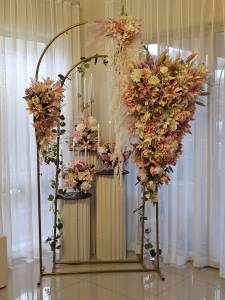 An elegant floral display featuring two interlocking gold metal arches. A large, cascading arrangement of peach lilies, white roses, and greenery hangs from the right side, while a smaller floral cluster sits on the left. In the foreground, white fluted pedestals hold additional flower arrangements and lit taper candles.
