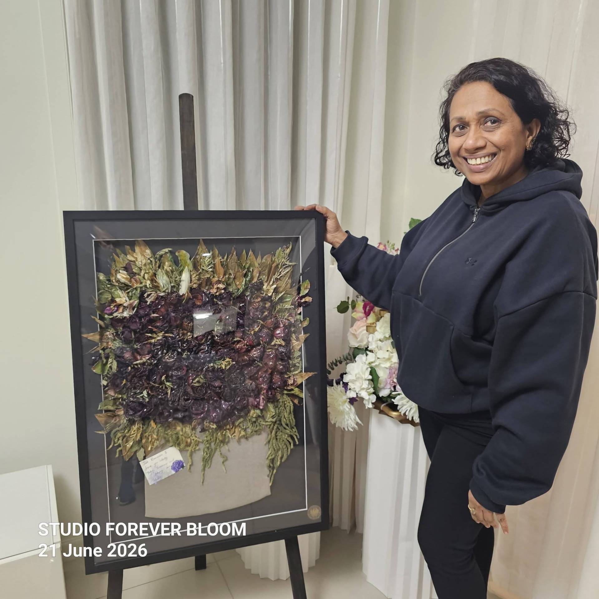 3D Pressed Flower Frame Preservation - Custom Wedding & Memorial Keepsake V1 A smiling floral artist showcasing a large black custom 3D pressed flower frame featuring preserved crimson roses and a personalized note card on an easel.