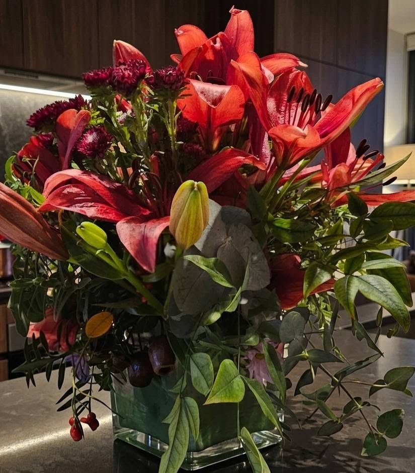 A luxury winter-themed floral display named WINTER LILIES Red Hybrid Arrangement V1 featuring large red lilies, dark burgundy blooms, and eucalyptus by Studio Forever Bloom.