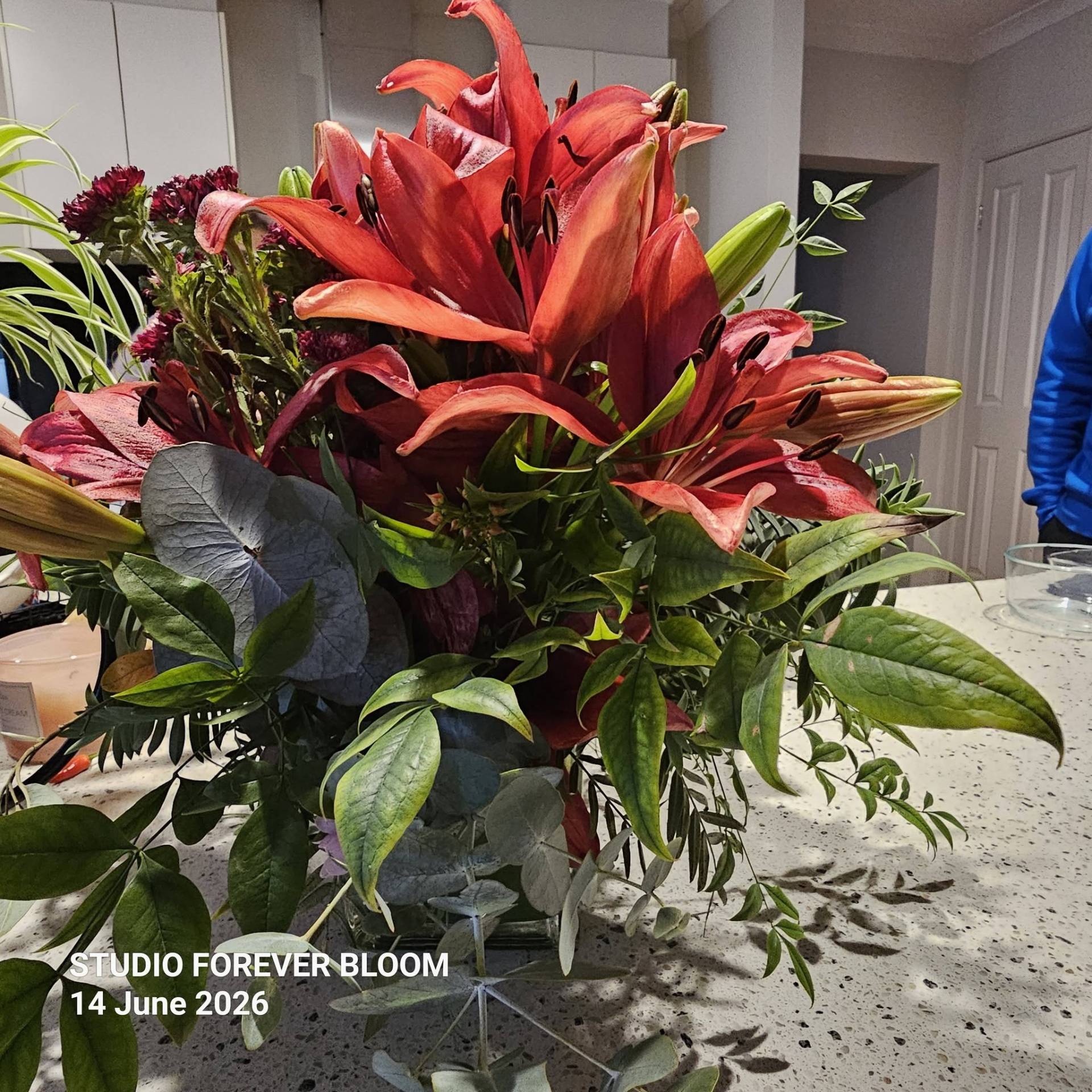 A luxury winter-themed floral display named WINTER LILIES Red Hybrid Arrangement V1 featuring large red lilies, dark burgundy blooms, and eucalyptus by Studio Forever Bloom.