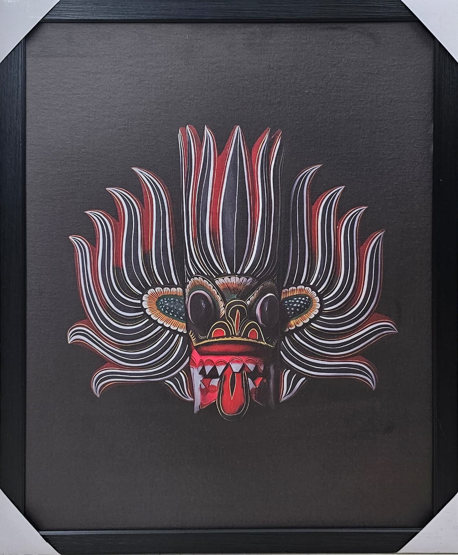 Framed 50x70 cm Traditional Sri Lankan Mask Art featuring intricate red and white patterns on a black background displayed on a wooden surface.